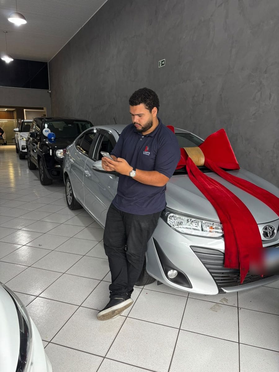 Gabriel Braga dentro da loja, encostado em um carro com laço vermelho, olhando o celular